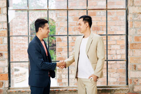 Happy young Asian businessman shaking hand of his smiling coworker in big loft office with brick wall and large windowsの写真素材