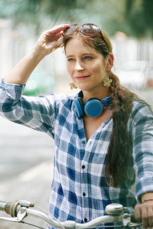 Portrait of pretty young smiling woman in plaid shirt sitting on bicycle and putting on sunglasses