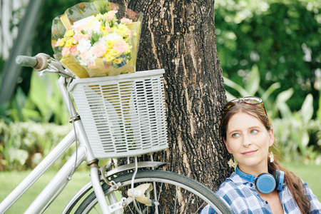Dreamy smiling young woman sitting under the tree in park and looking at flower bouquet in basket of her bicycleの写真素材