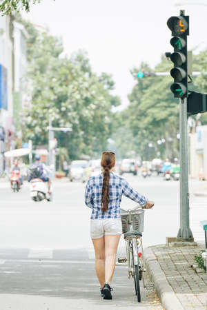 Rear view of young woman with long hair walking along city road with her bicycleの写真素材