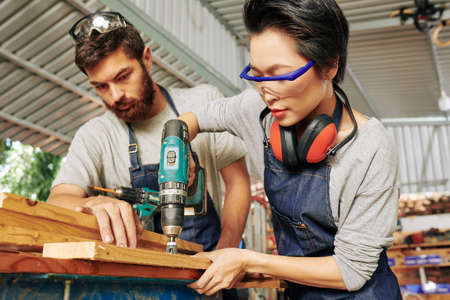 Serious bearded carpenter looking at his colleague in goggles drilling wooden plankの写真素材