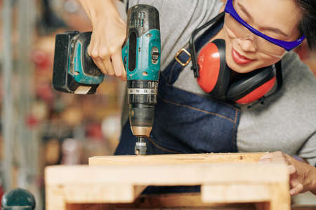 Smiling carpenter in plastic goggles drilling holes in wooden plankの写真素材