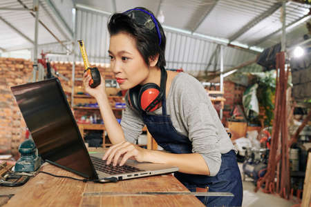 Asian female carpenter with tape measure checking 3D model of furniture piece of tablet computerの写真素材