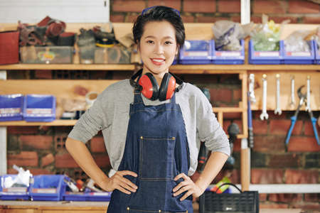 Portrait happy pretty young Vietnamese carpenter in denim overall standing with ear muiffs around her neck and hands on her hipsの写真素材