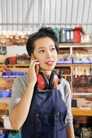 Pensive young Asian female carpenter in denim overall answering questions of client