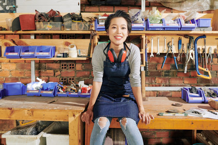 Happy pretty young Vietnamese female carpenter sitting on workbench in furniture workshopの写真素材