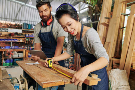 Positive female carpenter in denim apron measuring wooden plank with tape measureの写真素材