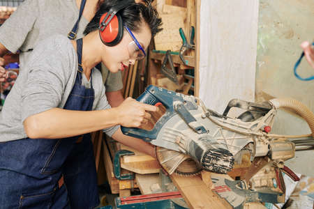 Professional young Asian female carpenter in ear muffs using circular saw when cutting pine wooden planksの写真素材