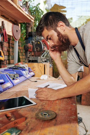 Serious bearded carpenter talking on phone with client and drawing sketch of furniture iteam on paper sheetの写真素材