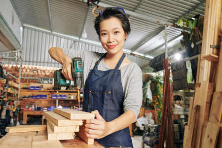 Portrait of pretty young Vietnamese carpenter using electric screwdriver when making furnuture at woodwork studioの写真素材