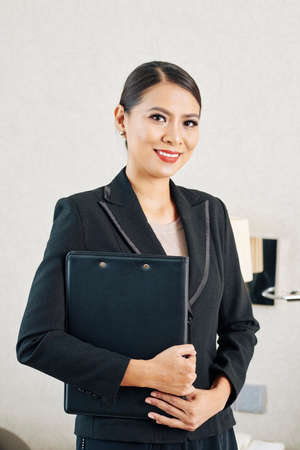 Portrait of Asian young businesswoman in formal black suit holding folder with documents and smiling at cameraの写真素材
