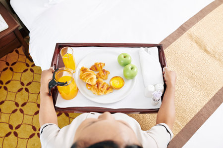 High view of young waitress standing and holding tray with croissants, juice and fruits she serving...