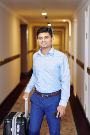Portrait of Indian young businessman with luggage standing in the corridor of the hotel and smiling...