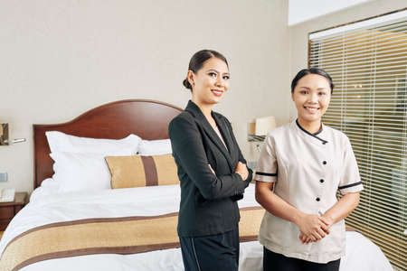 Portrait of Asian young manager in black suit standing together with Asian maid in uniform in the hotel room and they smiling at cameraの写真素材