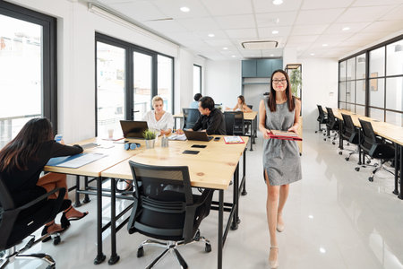 Young Asian businesswoman or secretary with folders walking along the office corridor with other...