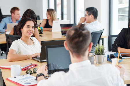 Young smiling businesswoman sitting at the table in front of laptop computer and listening to her colleagues during meeting at officeの写真素材