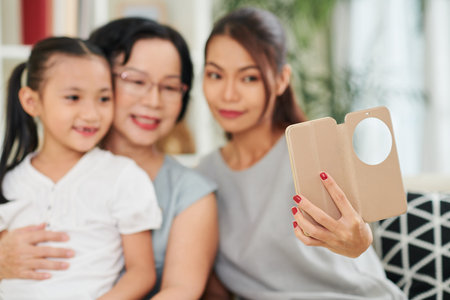 Young woman holding mobile phone and making selfie portrait of her mother and little daughter while...