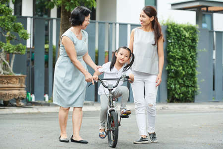 Happy Asian family mother and daughter talking to each other while teaching girl to ride on bicycleの写真素材