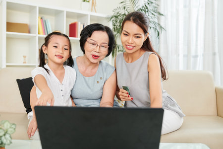 Asian family of three sitting on sofa together and communicating online using laptop computer at...