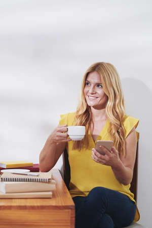 Pretty female student drinking cup of coffee and texting friends during short break after studying at homeの写真素材