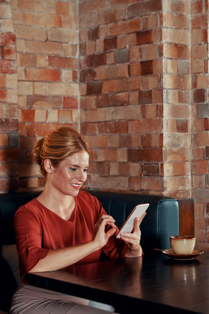 Pretty smiling Caucasian woman sitting at table in cafe and answering text messages from her friends...