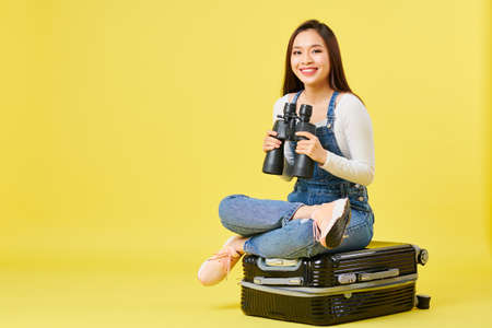 Happy beautiful young Asian woman in denim overall sitting on suitcase with binoculars and looking at cameraの写真素材
