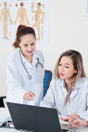 Pretty young Vietnamese doctor discussing information on laptop screen with her experienced coworker