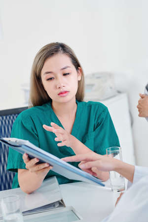 Serious young Asian nurse discussing medical history of patient with general practitionerの写真素材