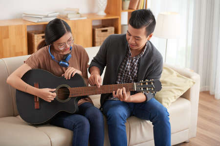Asian young man showing to his girlfriend how to play on guitar while they sitting on couch in the living room at homeの写真素材