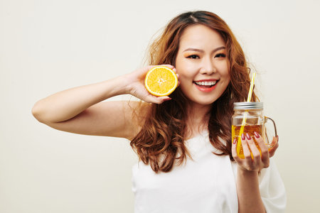 Portrait of Asian smiling young woman with red long curly hair holding orange fruit in one hand and...