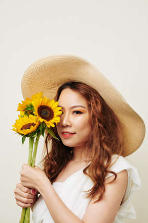 Portrait of red haired woman in hat holding bouquet of sunflowers near her face and looking at camera isolated on white backgroundの写真素材