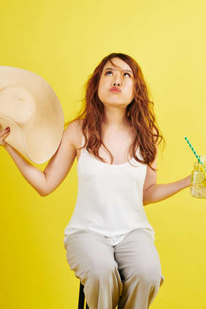 Asian fashion model sitting on chair with glass of juice and waving her hat because of heat she tired of shootingの写真素材