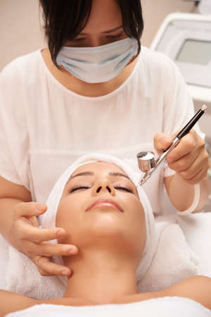 Asian cosmetologist in protective mask doing spa procedure with special equipment for young womans face while she lying with her eyes closedの写真素材