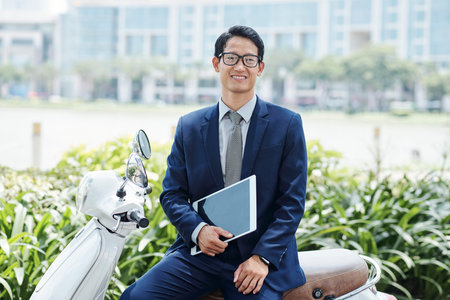 Portrait of young cheerful Asian businessman in glasses sitting on his scooter with digital tablet...