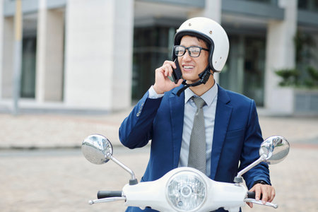 Portrait of young handsome Asian entrepreneur sitting on scooter and smiling when talking on phone