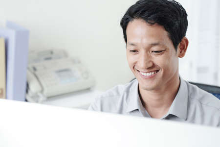 Happy handsome young Asian businessman reading document or e-mail on screen of computerの写真素材