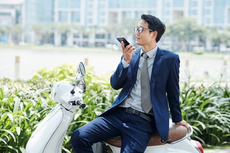Young handsome Vietnamese entrepreneur sitting on scooter and recording audio message for employee
