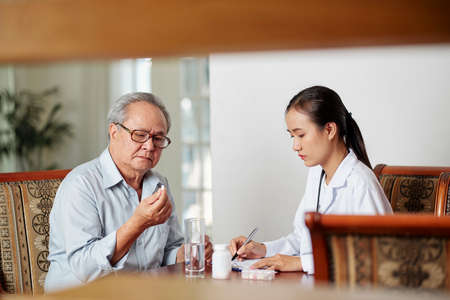 Asian female doctor sitting at the table and prescribing a medicine for treatment to her senior patient while he sitting opposite her and drinking pillの写真素材