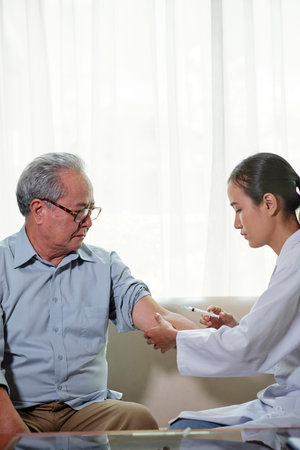 Young nurse in white coat sitting on sofa and make an injection in hand to her senior patient at...