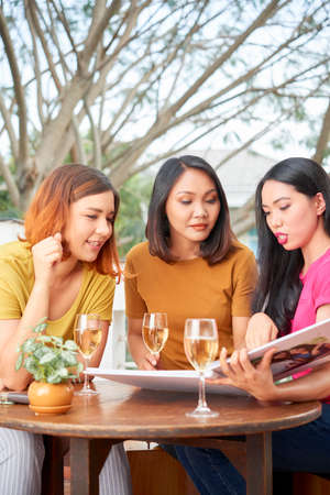 Three friends sitting at the table looking photos in photo album and remembering happy moments together while drinking champagne in cafeの写真素材