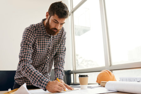 Serious bearded architect in casual clothing working at his workplace he standing and drawing...