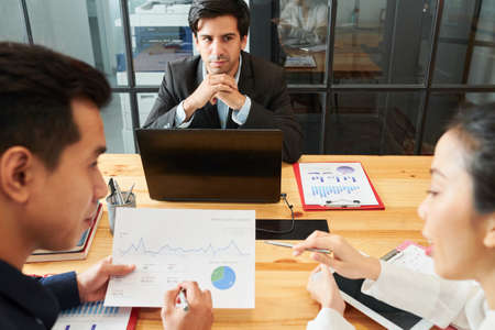 Young businessman sitting at the table in front of laptop and listening to his partners while they discussing financial report during meeting at officeの写真素材