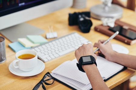 Business person checking notifications on smartwatch when writing down plans and ideas in diary,...