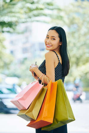 Portrait of Asian stylish girl with paper shopping bags smiling at camera while standing outdoors in the cityの写真素材