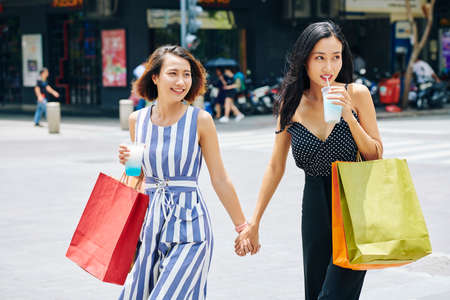 Two Asian young women with shopping bags holding hands and drinking cocktails while walking along the street in big cityの写真素材