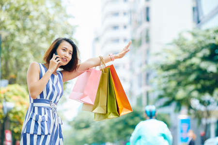 Beautiful Asian woman in dress with shopping bags has a conversation on mobile phone while catching a taxi outdoors in the cityの写真素材