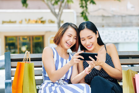 Two Asian young women resting on bench after shopping and watching funny photos on mobile phone together in the cityの写真素材
