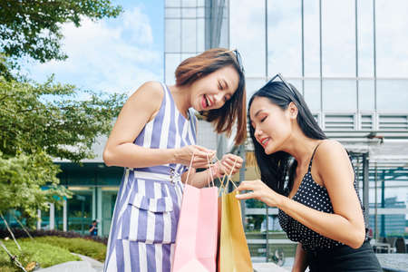 Happy Asian woman showing her purchases after shopping to her friend while they meeting in the cityの写真素材