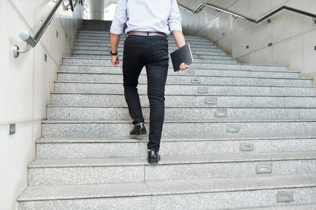 Rear view of young man with digital tablet in hand walking up the stairs and hurrying to office