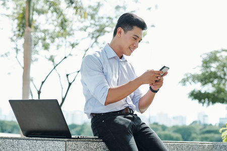Smiling Asian digital nomad sitting outdoors next to his laptop and checking text messages in...